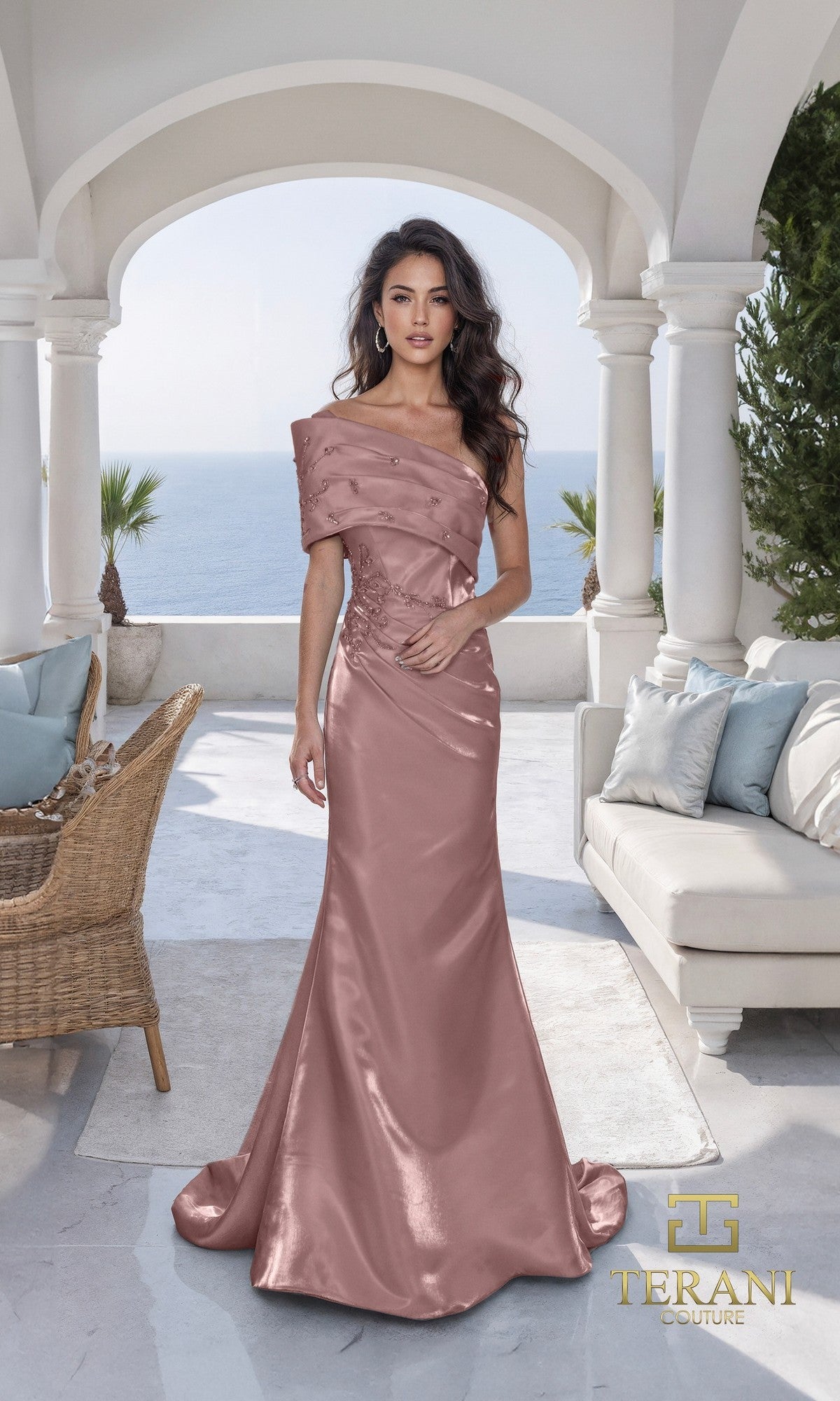 A woman in an espresso, off-shoulder satin floor-length dress (Long Formal Dress: Terani 261E6227) stands on a terrace with white arches, overlooking the sea and palm trees. The